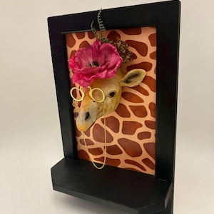 May include: A wall-mounted display featuring a sculpted giraffe head wearing gold-rimmed spectacles with a delicate chain. The giraffe is adorned with a large pink flower and sits against a background with a brown and tan giraffe print pattern.