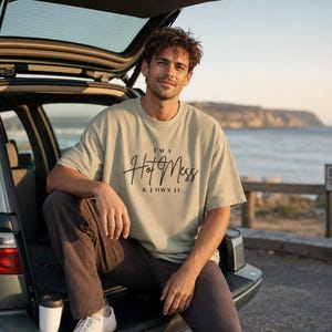 Op de afbeelding: Een saliegroen t-shirt met korte mouwen en de zwarte tekst "I'M A Hot Mess & I OWN IT." op de borst gedrukt. Het kledingstuk heeft een ronde hals en een relaxte pasvorm, gemaakt van een effen stof met een eenvoudig en strak ontwerp.