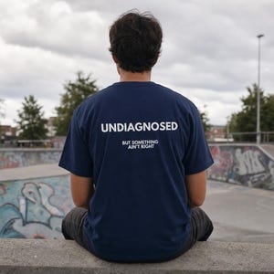 Op de afbeelding: Een marineblauw t-shirt met korte mouwen en witte tekst op de achterkant met de tekst UNDIAGNOSED in grote hoofdletters, gevolgd door de kleinere tekst BUT SOMETHING AIN'T RIGHT gecentreerd onder het hoofdwoord.