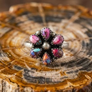 Könnte beinhalten: Ein Silberring mit einer zentralen Metallkugel, umgeben von sechs tropfenförmigen Steinen in den Farben Pink, Violett, Orange und Türkis. Die Steine sind in einzelnen Silberfassungen in einem Blumenmuster auf dem Ringband angeordnet.