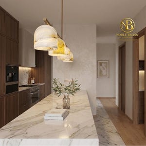 May include: A linear pendant light fixture featuring four dome-shaped shades with a marbled white and gold finish, suspended by a brass rod above a kitchen island. The Noble Brass logo is visible in the upper right corner of the image.