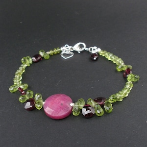 Può includere: Un braccialetto d'argento con pietre preziose verdi e viola. Il braccialetto presenta una grande pietra preziosa rotonda rosa al centro.