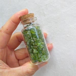 Puede incluir: Una pequeña botella de vidrio con un tapón de corcho llena de cristales de peridoto verdes.