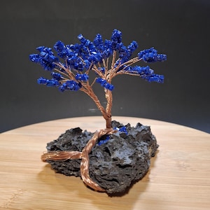 Op de afbeelding: Een handgemaakte draadboomsculptuur met een koperkleurige stam en takken. Het gebladerte van de boom is gemaakt van levendige blauwe edelstenen. De boom is gemonteerd op een donkere, getextureerde rotsbasis.