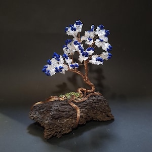 May include: A handcrafted wire tree sculpture with a copper-colored trunk and branches. The tree's foliage is composed of blue and clear crystal beads. The tree is mounted on a dark, textured, stone base, creating a unique decorative piece.