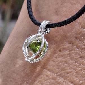 Op de afbeelding: Een zilveren hangerketting met een kooi-achtig ontwerp met een vierkante, lichtgroene edelsteen. De hanger hangt aan een zwart koord. De edelsteen is een levendig, opvallend groen.