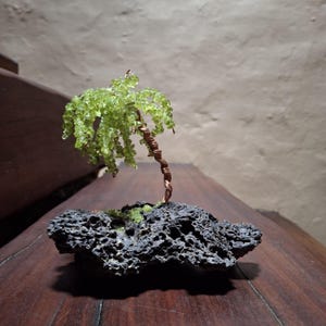 May include: A decorative sculpture of a tree featuring a twisted copper wire trunk and branches adorned with small, translucent light green gemstone chips. The tree is anchored to a dark, porous, textured volcanic rock base.