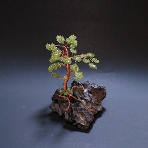 Op de afbeelding: Een handgemaakte draadboomsculptuur met een koperen stam en takken, versierd met groene edelsteenbladeren. De boom is geworteld in een donkere, getextureerde rotsbasis, waardoor een miniatuur bonsai-achtige esthetiek ontstaat.