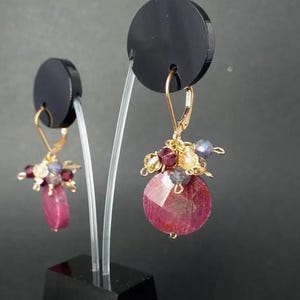 Può includere: Un paio di orecchini dorati con chiusura a monachella, caratterizzati da un grappolo di piccole perline sfaccettate multicolori nei toni del rosso, giallo e blu, con una grande pietra preziosa rotonda e sfaccettata rosso rosato alla base.