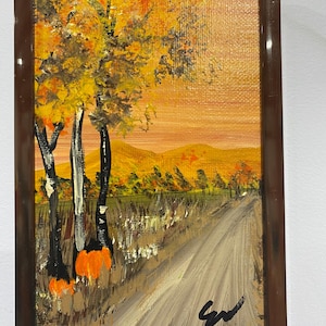 May include: A rectangular framed painting depicting an autumn landscape with a dirt path leading towards distant golden hills. The scene features trees with yellow and orange foliage, tall grasses, and a bright orange sky. A black signature is in the corner.