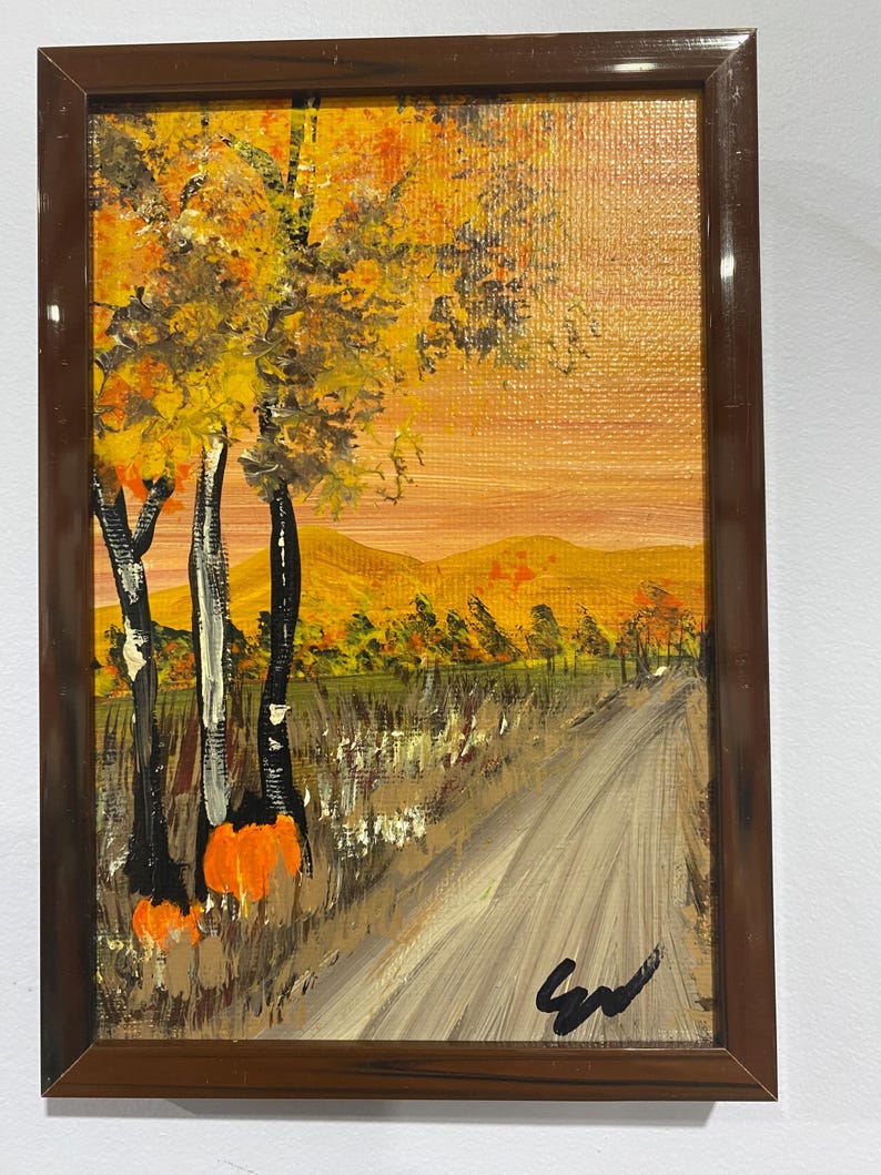 May include: A rectangular framed painting depicting an autumn landscape with a dirt path leading toward distant golden hills. The scene features trees with yellow and orange foliage, tall grasses, and a bright orange sky. A black signature is in the corner.