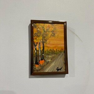 May include: A rectangular framed painting depicting an autumn landscape with a dirt path leading toward distant hills. The scene features tall trees with yellow and orange foliage, dark tree trunks, and a warm, golden sky. A black signature is in the corner.