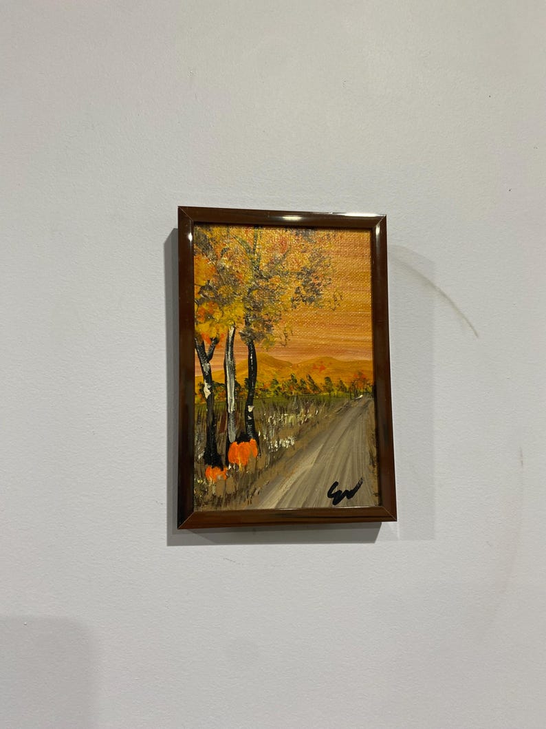 May include: A rectangular framed painting depicting an autumn landscape with a dirt path leading toward distant hills. The scene features tall trees with yellow and orange foliage, dark tree trunks, and a warm, golden sky. A black signature is in the corner.