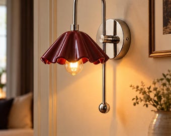 May include: A wall-mounted lamp featuring a polished silver-tone metal arm and a glossy, deep red, scalloped ceramic shade. The fixture includes a round wall plate and a vertical rod ending in a spherical finial, with a warm light bulb visible.