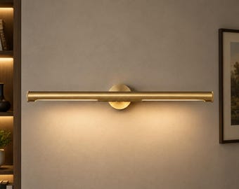 May include: A horizontal wall-mounted light fixture with a brushed brass finish. The cylindrical lamp features a central circular mounting plate and emits a warm, downward-facing glow along its length, illuminating the wall surface below.