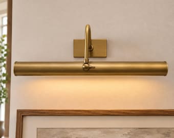 May include: A wall-mounted picture light with a brushed brass finish. The fixture features a rectangular backplate, a single curved arm, and a long cylindrical tube housing the light source, which emits a warm glow downward onto the wall.