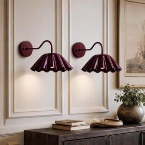 May include: A pair of wall-mounted sconces featuring deep burgundy, scalloped, fluted shades. Each lamp has a matching circular wall plate and a curved arm that extends outward, directing light downward from the wide, pleated metal shade design.