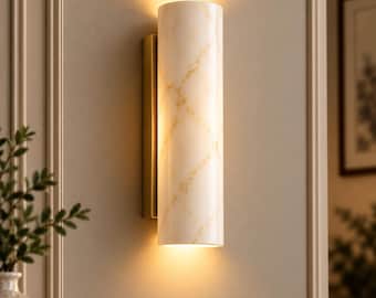May include: A cylindrical wall sconce featuring a white marble shade with subtle gold veining, mounted on a rectangular brass-toned base. The fixture emits a warm, soft glow from both the top and bottom, illuminating the wall surface behind it.