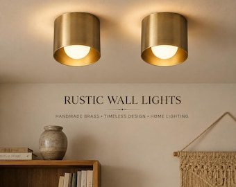 May include: Two cylindrical brass wall lights mounted on a ceiling, each featuring a warm glowing bulb. The text RUSTIC WALL LIGHTS, HANDMADE BRASS, TIMELESS DESIGN, HOME LIGHTING is printed on the wall below the lights, above a wooden shelf.