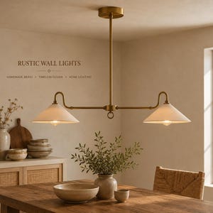 May include: A brass chandelier with two conical off-white shades suspended from a central rod. The fixture features a horizontal arm design with a small ring detail at the centre. Text on the wall reads RUSTIC WALL LIGHTS, HANDMADE BRASS, TIMELESS DESIGN, HOME LIGHTING.