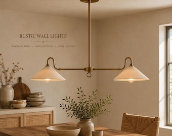 May include: A brass chandelier with two conical off-white shades suspended from a central rod. The fixture features a horizontal arm design with a small ring detail at the center. Text on the wall reads RUSTIC WALL LIGHTS, HANDMADE BRASS, TIMELESS DESIGN, HOME LIGHTING.