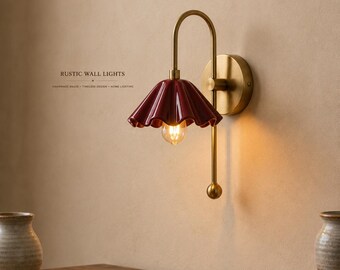 May include: A wall lamp featuring a curved brass arm and a deep red, fluted ceramic shade. The fixture includes a circular brass wall mount and a small brass sphere at the base of the arm. Text on the wall reads RUSTIC WALL LIGHTS.