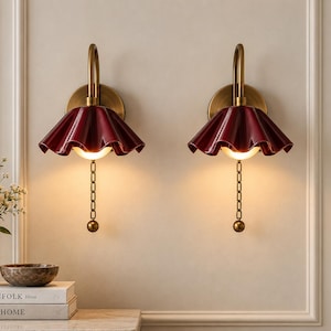 May include: A pair of wall sconces featuring a deep red, ruffled, bell-shaped shade attached to a curved brass arm. Each fixture includes a circular brass wall mount, a glowing light bulb, and a vertical brass pull chain with a spherical end.