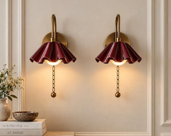May include: A pair of wall sconces featuring a deep red, ruffled, bell-shaped shade attached to a curved brass arm. Each fixture includes a circular brass wall mount, a glowing light bulb, and a vertical brass pull chain with a spherical end.