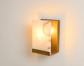 May include: A wall sconce featuring a rectangular white marble panel with grey veining, held by a brushed brass metal frame. The fixture emits a warm, soft glow from behind the marble, casting light onto the wall and highlighting the stone texture.