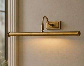 May include: A wall-mounted picture light fixture with a brushed brass finish. The design features a rectangular base plate, a curved arm, and a long, horizontal cylindrical light bar that emits a warm, downward-facing glow onto the wall surface.