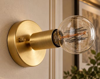 May include: A wall-mounted light fixture with a circular brushed brass base and a cylindrical socket. A large, clear globe-shaped light bulb with visible internal filaments is screwed into the socket. A small brass knob is located on the side.