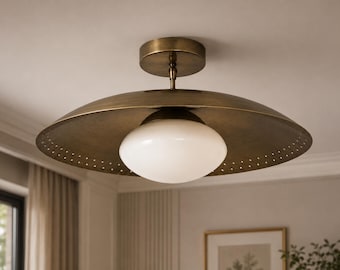 May include: A semi-flush mount ceiling light fixture featuring a wide, circular, antique brass-finished metal shade with a perforated edge. A central, spherical, opaque white glass globe sits beneath the shade, attached to a matching brass canopy.