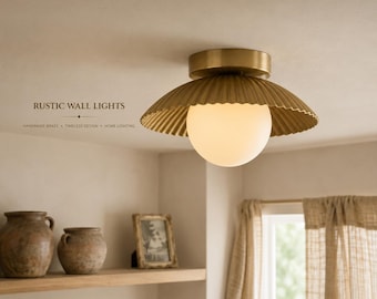 May include: A brass ceiling light fixture featuring a wide, pleated, metallic shade and a central spherical white glass globe. The text RUSTIC WALL LIGHTS HANDMADE BRASS TIMELESS DESIGN HOME LIGHTING is printed on the left side of the image.