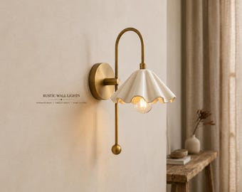 May include: A wall lamp featuring a brass arm with a circular wall mount and a downward-facing, scalloped white ceramic shade. The fixture includes a small spherical finial at the bottom of the arm and a single light bulb. Text reads RUSTIC WALL LIGHTS.