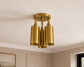 May include: A brass ceiling light fixture featuring three cylindrical shades attached to a central circular mount. Each shade is connected by a swivel joint, allowing for adjustable positioning of the light heads in a downward direction.