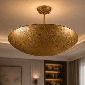 May include: A large, circular, gold-toned metal pendant light fixture suspended from the ceiling by a thin rod. The dome-shaped shade features an intricate, embossed pattern across its entire surface and a matching textured circular canopy.