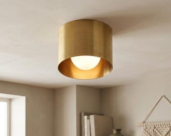May include: A cylindrical flush mount ceiling light fixture with a brushed brass finish. The fixture features a circular metal housing that surrounds a central, spherical white glass globe bulb, emitting a soft, warm glow from within the shade.