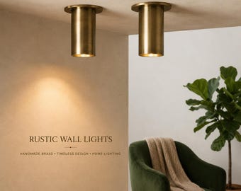 May include: Two cylindrical brass wall lights mounted on a ceiling, featuring a warm metallic finish. The text RUSTIC WALL LIGHTS, HANDMADE BRASS, TIMELESS DESIGN, HOME LIGHTING is printed on the wall below the lights, which cast a soft glow.