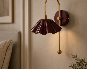 May include: A wall-mounted lamp featuring a deep burgundy, pleated, bell-shaped shade and a circular wall plate in the same color. The fixture is supported by a curved, brushed gold-tone metal arm with a matching gold-tone ball finial at the base.
