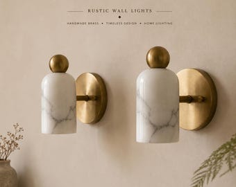 May include: Two wall lights featuring cylindrical white marble shades with grey veining, topped with spherical brass finials. Each light is mounted on a circular brass wall plate. Text above reads: RUSTIC WALL LIGHTS, HANDMADE BRASS, TIMELESS DESIGN, HOME LIGHTING.