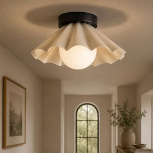 May include: A flush mount ceiling light featuring a matte black circular base and a wide, cream-colored, ruffled ceramic shade. A central, spherical, frosted white glass bulb hangs downward, emitting a soft, diffused glow from the fixture.