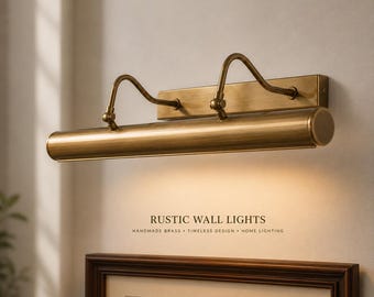 May include: A brass picture light fixture with a horizontal cylindrical lamp head and two curved adjustable arms mounted on a rectangular wall plate. The text RUSTIC WALL LIGHTS HANDMADE BRASS TIMELESS DESIGN HOME LIGHTING is visible below.