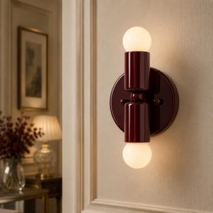 May include: A wall sconce featuring a circular dark burgundy base and a vertical cylindrical body in the same deep red finish. The fixture holds two spherical white glass bulbs, one positioned at the top and one at the bottom, emitting light.