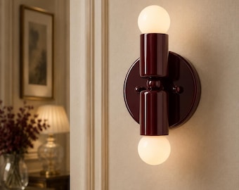 May include: A wall sconce featuring a circular dark burgundy base and a vertical cylindrical body in the same deep red finish. The fixture holds two spherical white glass bulbs, one positioned at the top and one at the bottom, emitting light.