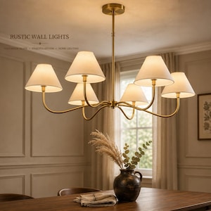 May include: A six-light chandelier featuring a brass finish frame with curved arms and off-white fabric shades. The fixture is suspended from a central rod and ceiling canopy. Text in the image reads RUSTIC WALL LIGHTS HANDMADE BRASS TIMELESS DESIGN HOME LIGHTING.