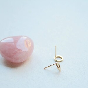 May include: A pair of small gold circle stud earrings with thin posts, positioned next to a smooth, polished, heart-shaped rose quartz stone. The earrings feature a simple open circular design, and the stone has a soft, translucent pink hue.