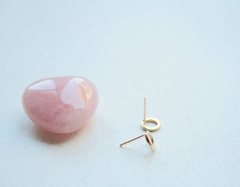 May include: A pair of small gold circle stud earrings with thin posts, positioned next to a smooth, polished, heart-shaped rose quartz stone. The earrings feature a simple open circular design, and the stone has a soft, translucent pink hue.
