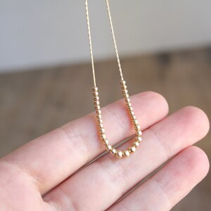 Pu&ograve; includere: Una delicata collana color oro caratterizzata da una catenina sottile decorata con una serie di piccole perline metalliche rotonde disposte a forma di U al centro. Le perline sono uniformi per dimensioni e colore, creando un aspetto raffinato.