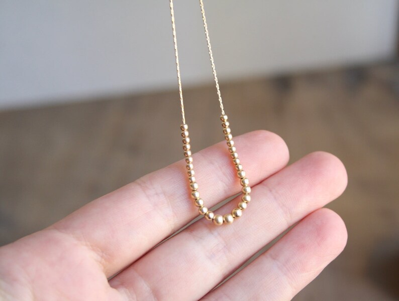 Pu&ograve; includere: Una delicata collana color oro caratterizzata da una catenina sottile decorata con una serie di piccole perline metalliche rotonde disposte a forma di U al centro. Le perline sono uniformi per dimensioni e colore, creando un aspetto raffinato.