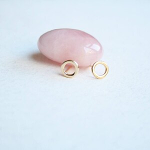 May include: A pair of small, circular gold stud earrings featuring a simple open-ring design. The earrings are positioned in front of a smooth, polished, pale pink rose quartz stone, highlighting the contrast between the metallic gold and the stone.